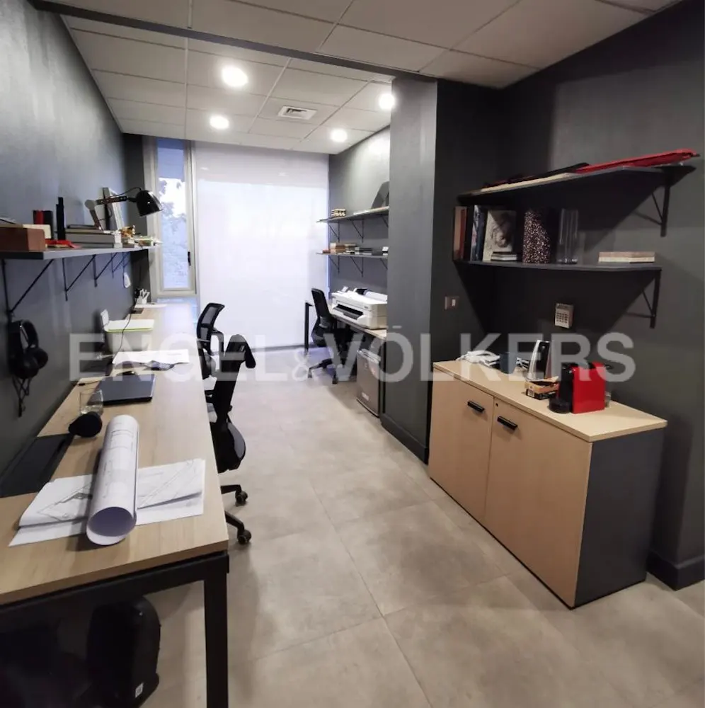 Office space with desks, chairs, and shelves. The walls are gray, and the floor is tiled. There is a printer and a cabinet in the background.