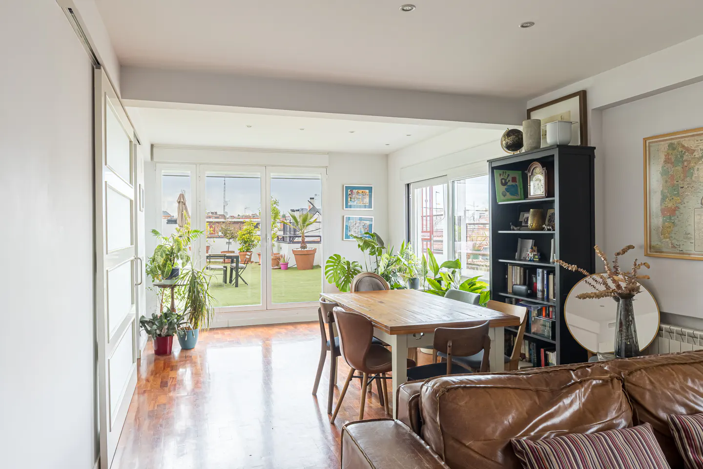 Bright living room with wood floors, a dining table, and a black bookshelf. Sliding glass doors lead to a rooftop patio with plants and furniture.
