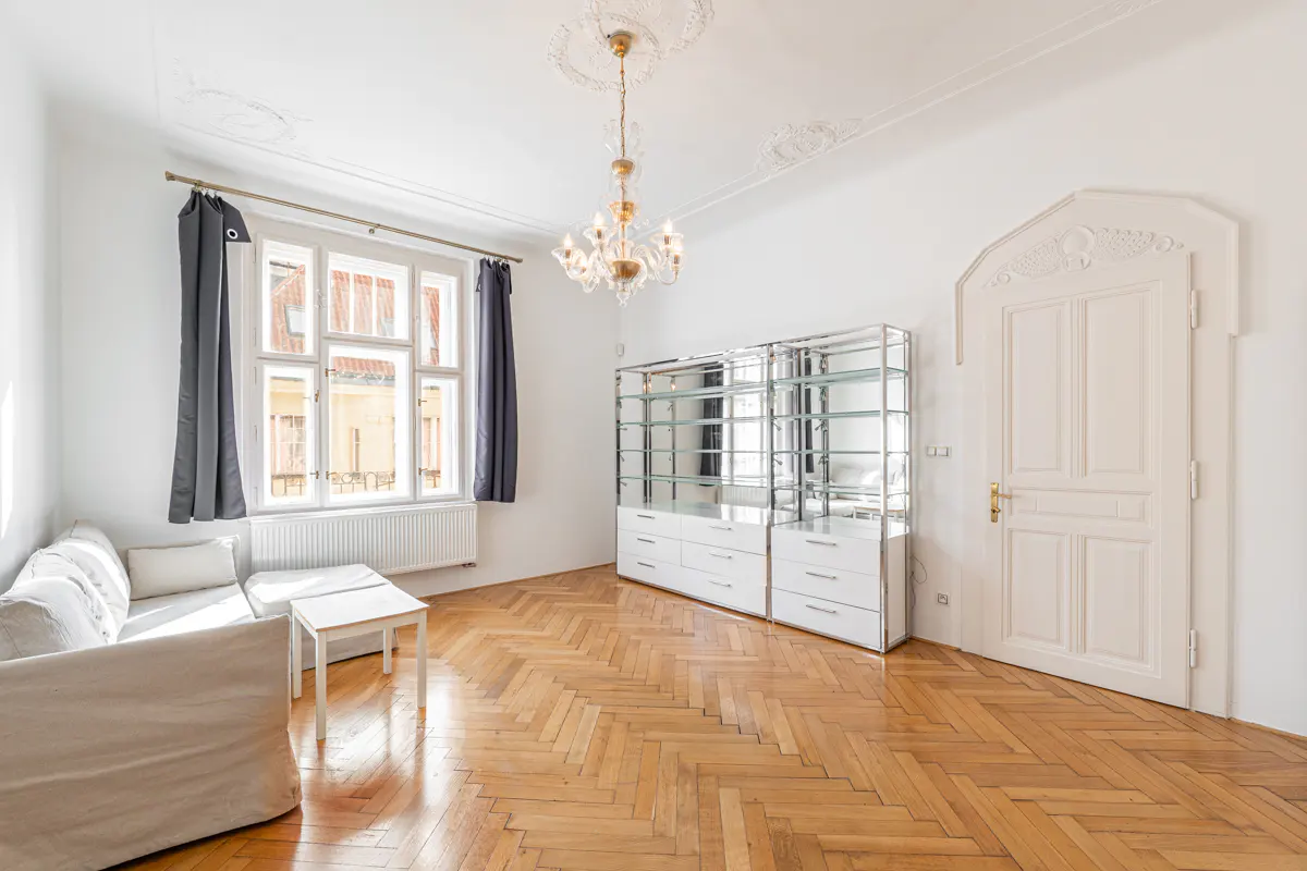 Bright living room with herringbone wood floors, white walls, and a crystal chandelier. A white sofa sits near a window with gray curtains.
