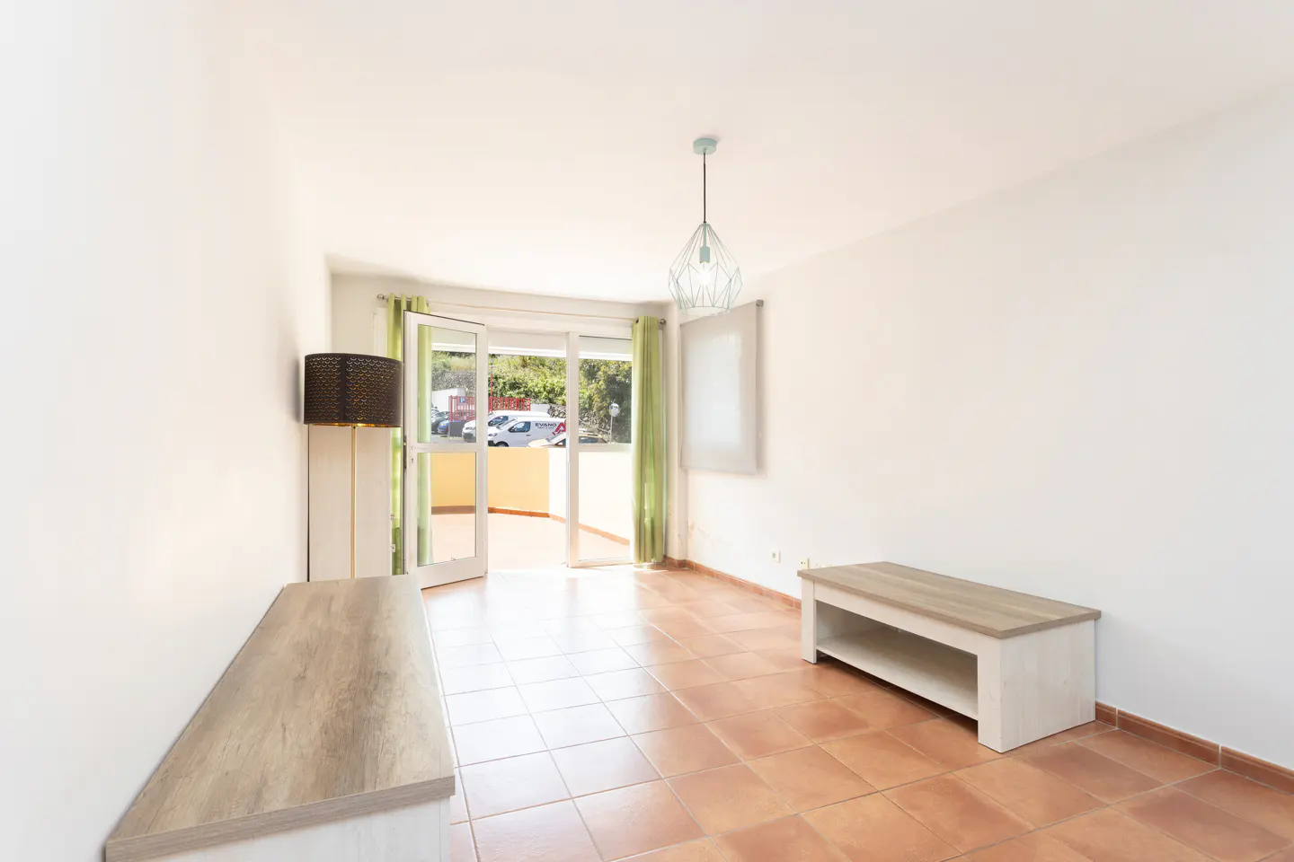 Bright, empty room with terracotta tile floor, white walls, and open balcony doors. A light fixture hangs from the ceiling. Two light wood consoles sit against the walls.