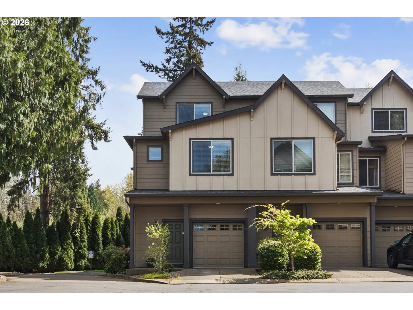 A three-story townhouse with a gray roof, beige siding, and a brown garage door. Green trees are in the background.