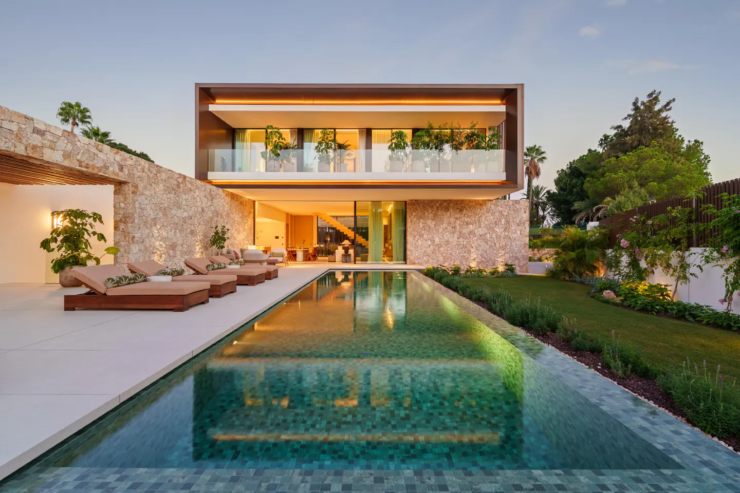 Modern two-story home with a pool. The house has a stone wall, a balcony with plants, and lounge chairs by the pool.