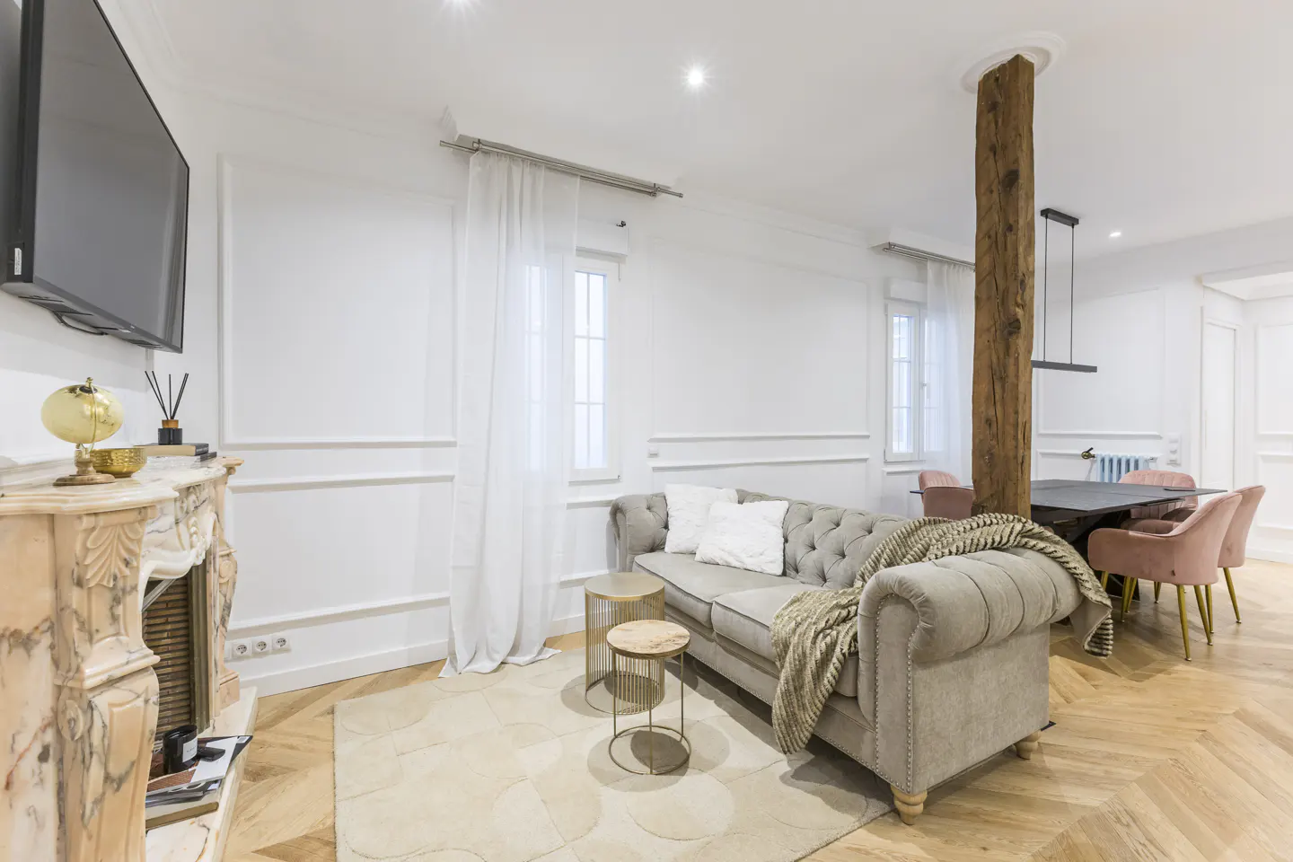 Bright living room with herringbone wood floors, a gray sofa, and a marble fireplace. A rustic wood column separates the living and dining areas.