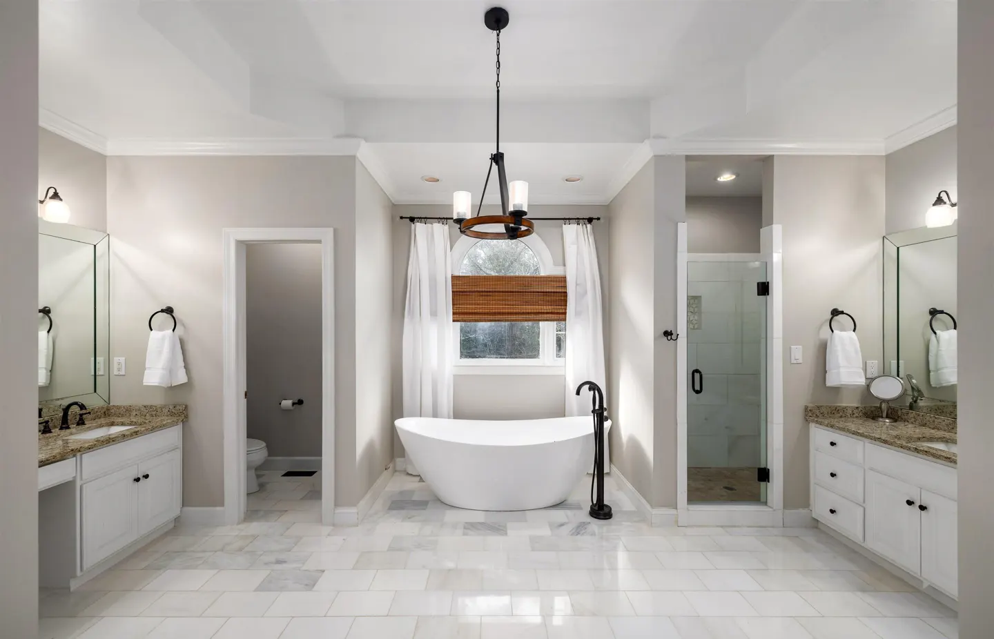 Bright bathroom with white marble floors, double vanities, a freestanding tub under a window, and a glass-enclosed shower.