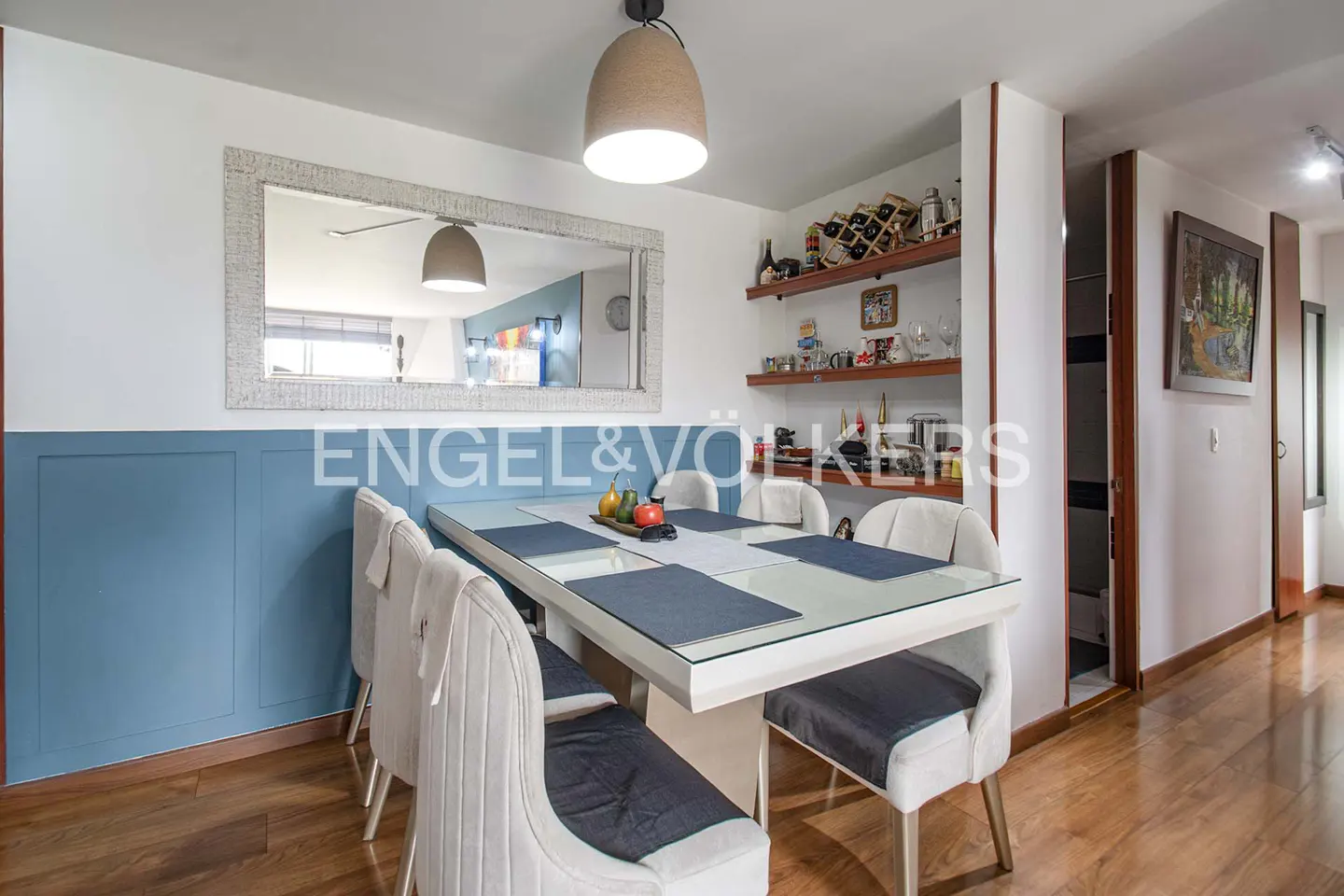 Dining room with a white table, six chairs, blue wall paneling, and wooden floors. Shelves with wine bottles and decor are on the wall.