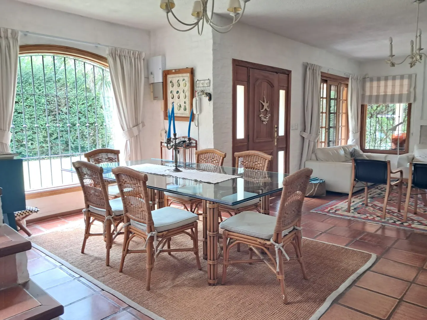 Dining room with a glass table, six wicker chairs, and a jute rug on a tiled floor, leading to a living area.