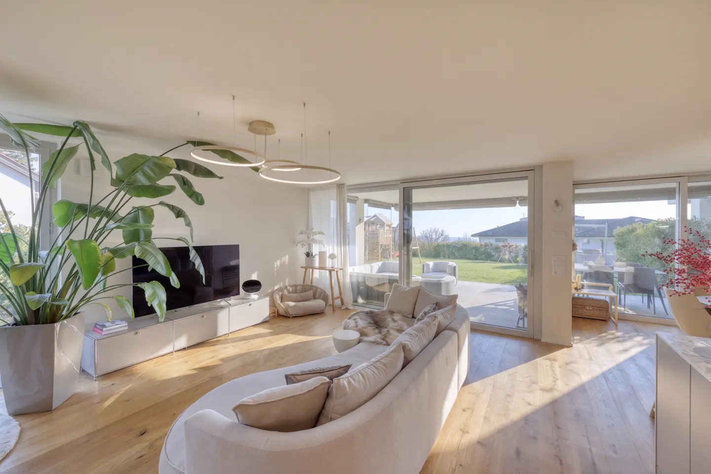 Bright living room with a curved white sofa, wood floors, and a large potted plant. Sliding glass doors open to a green lawn.