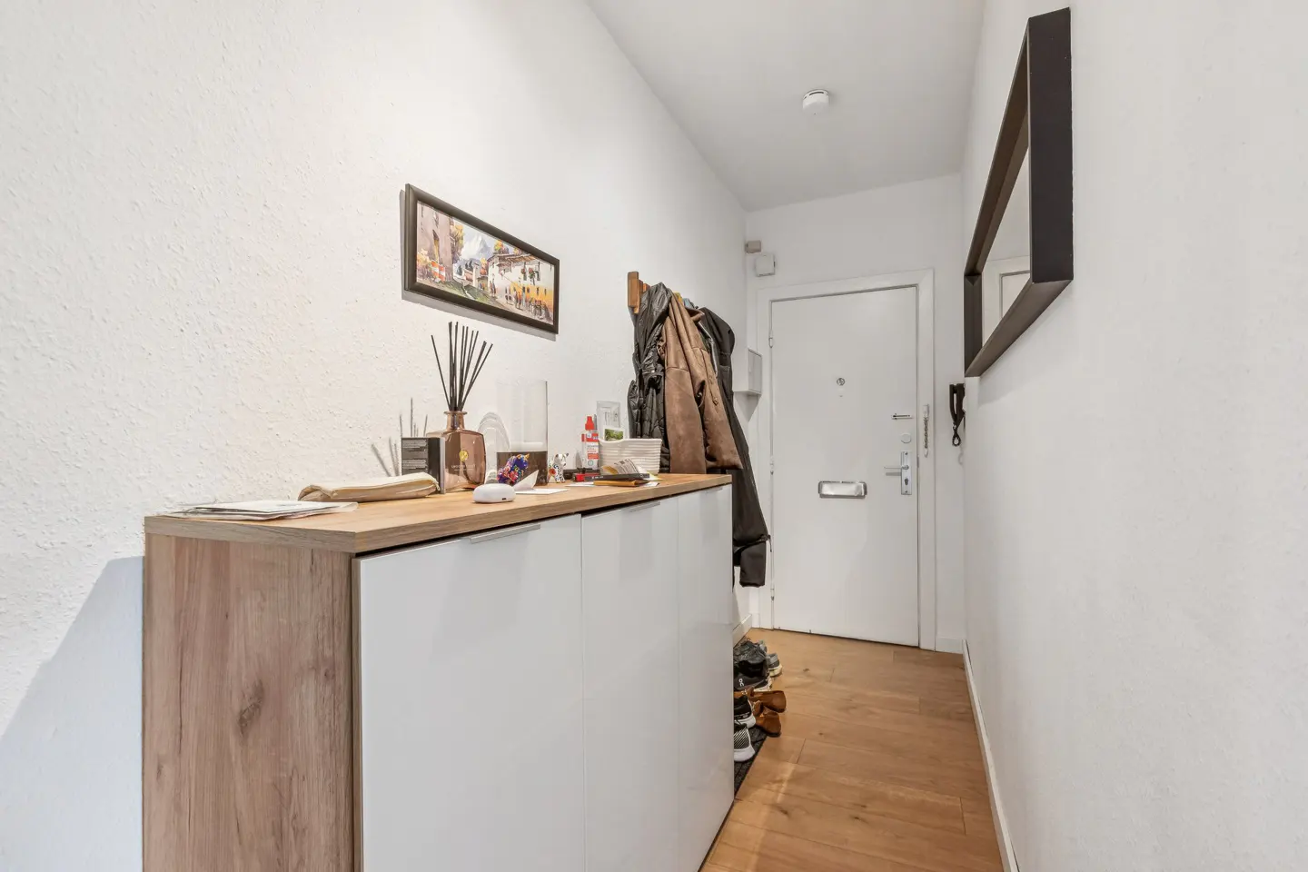 Hallway with white walls, wood floors, and a light wood and white cabinet. A framed picture hangs above the cabinet. A white door is at the end of the hall.