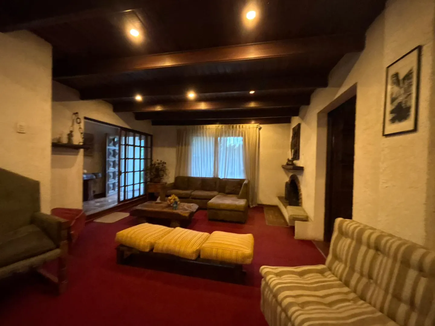Living room with red carpet, brown wood ceiling beams, and a fireplace. Couches and a coffee table furnish the space.