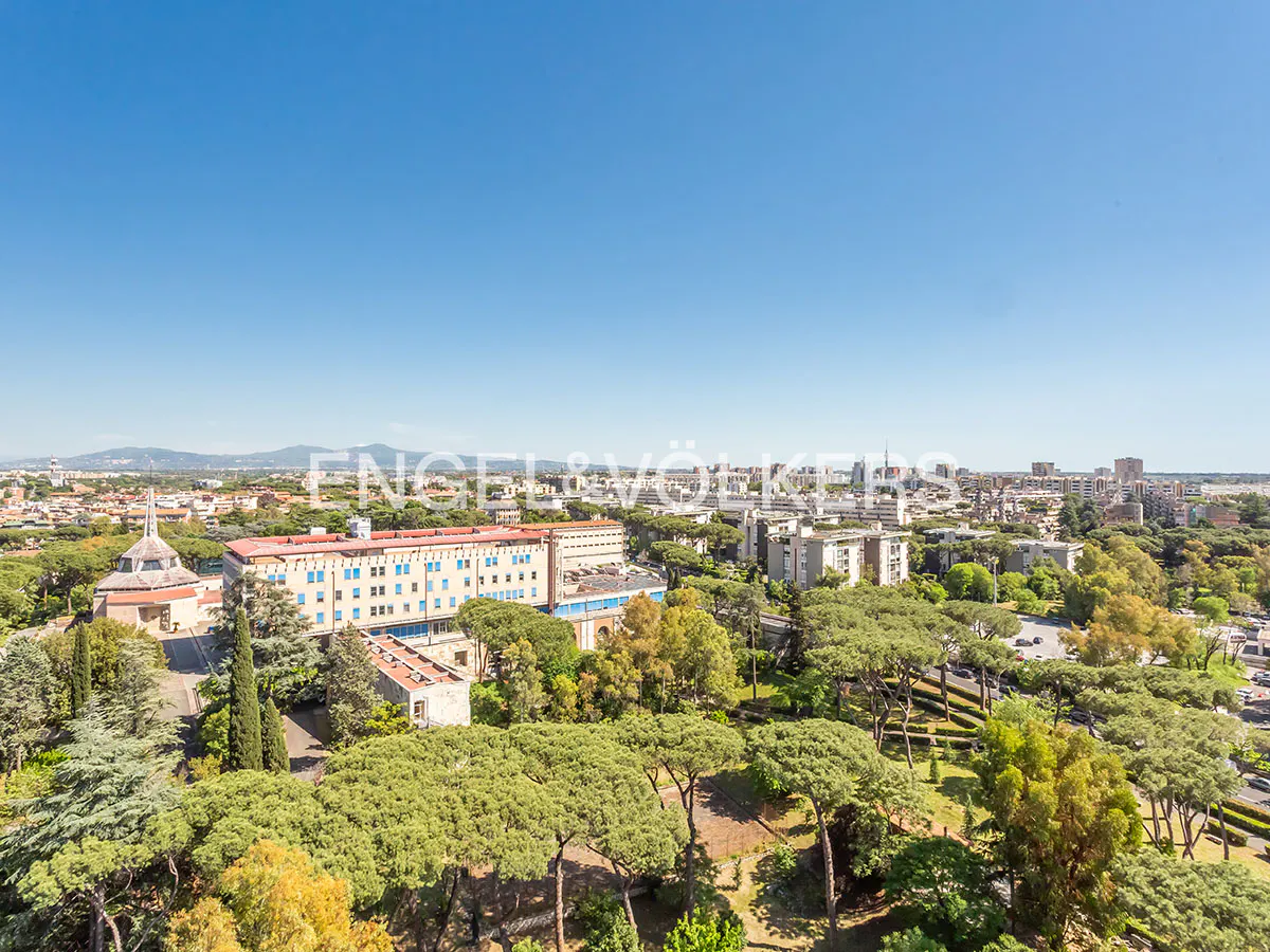 Cityscape view with green trees, buildings, and blue sky. A church steeple and mountains are visible in the distance.