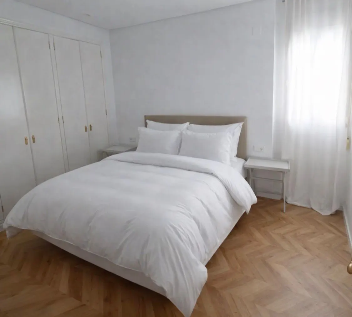 Bright bedroom with white bedding, a tan headboard, and white walls. A window with sheer curtains is on the right.