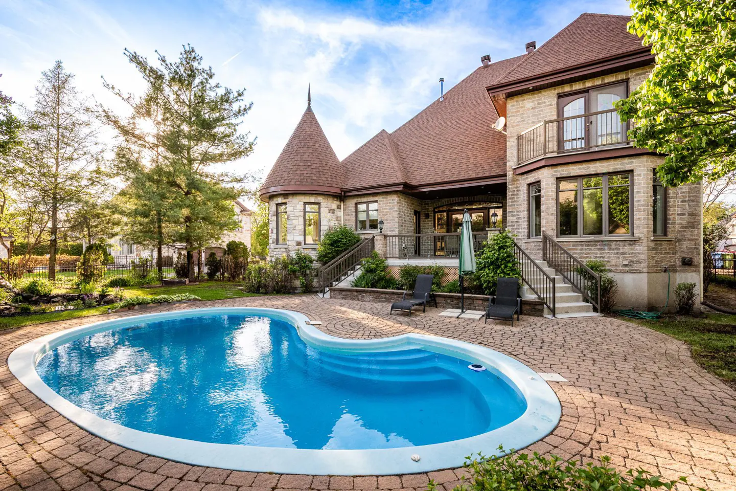 Backyard view of a large stone house with a kidney-shaped blue pool, brick patio, lounge chairs, and green trees under a blue sky.
