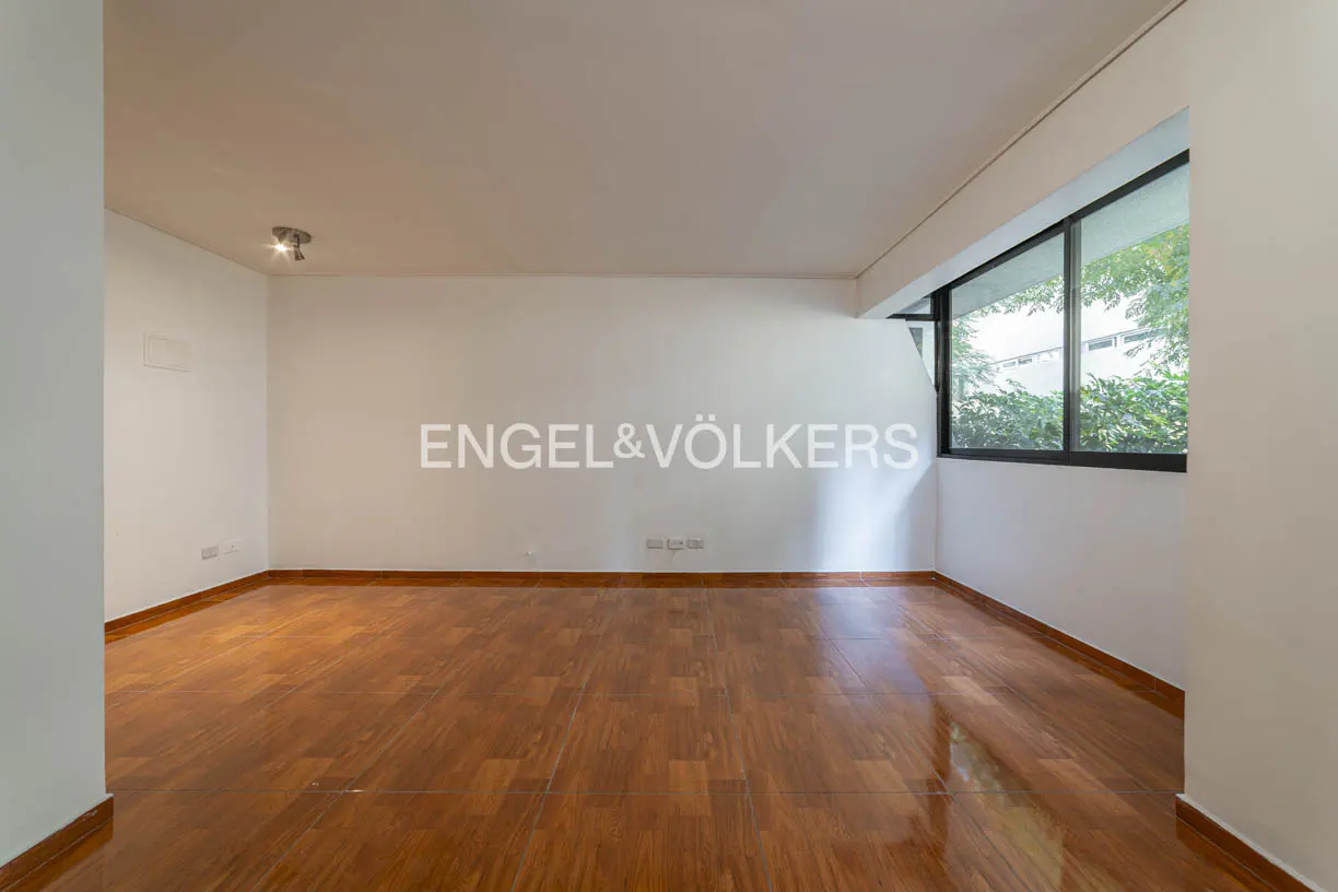 Empty room with hardwood floors, white walls, and a large window showing green trees. Engel & Völkers logo on the wall.