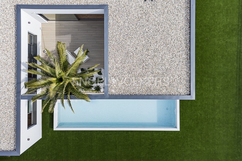 Aerial view of a modern house with a pool, gravel roof, and a palm tree on a wooden deck. Green lawn surrounds the property.
