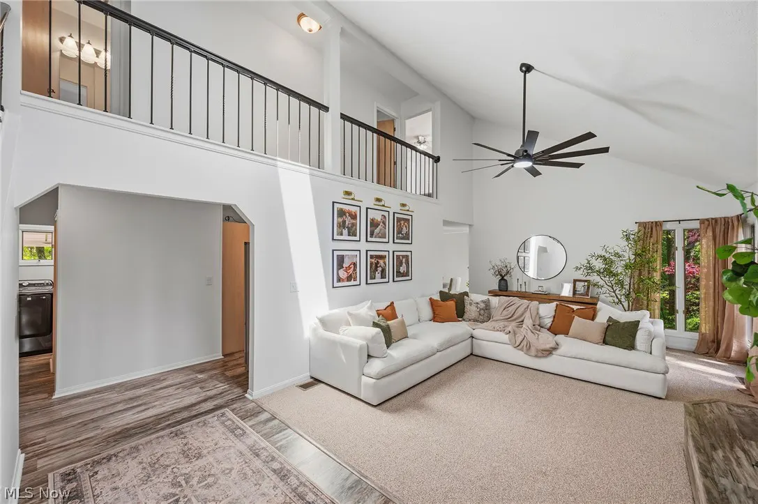 Bright living room with white walls, beige carpet, and a large white sectional sofa with colorful pillows. A black ceiling fan hangs above.