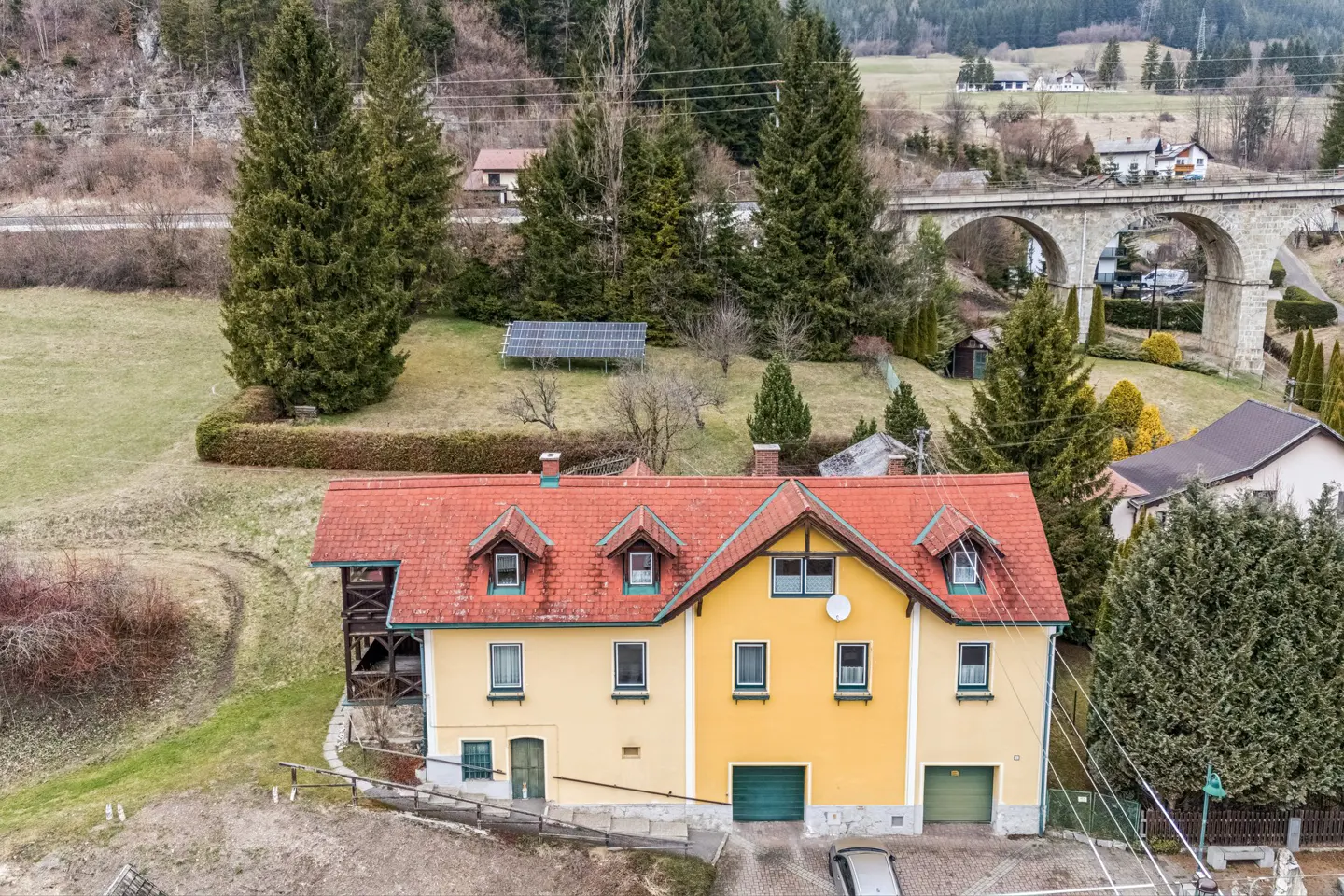 Aerial view of a yellow house with a red roof, green garage doors, and a stone arch bridge in the background.