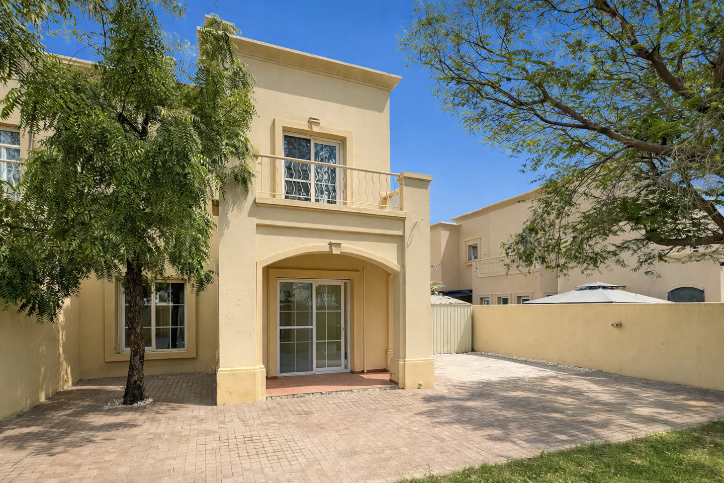 Two-story beige house with a balcony and sliding glass doors, a brick patio, and trees under a blue sky.