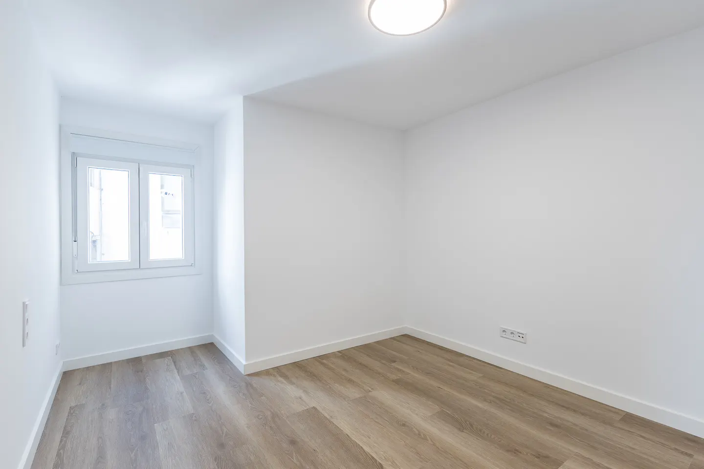 Bright, empty room with white walls, light wood floors, and a window with white trim. A flush ceiling light provides illumination.