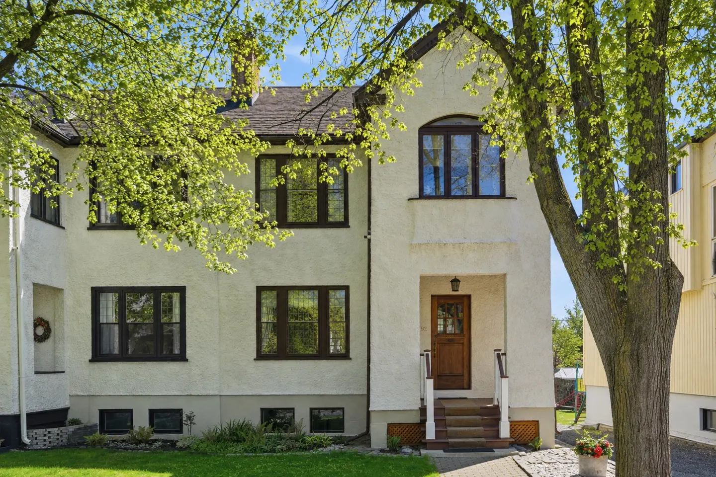 Two-story cream stucco house with dark brown window frames, a wooden front door, and lush green trees in the front yard.