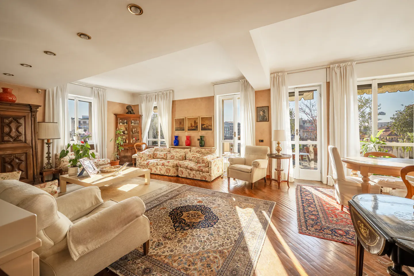 Bright living room with hardwood floors, floral sofa, cream armchairs, and ornate rugs. Sunlight streams through white-curtained windows.