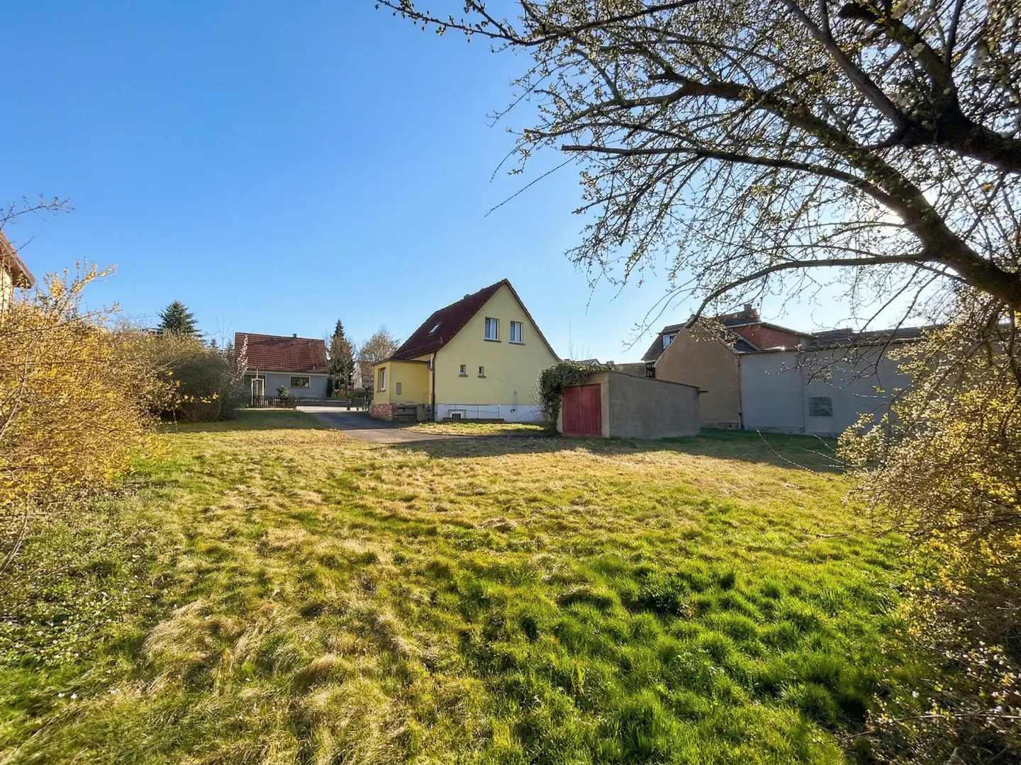 A yellow house with a red roof sits behind a grassy lawn under a blue sky. A tree is on the right.