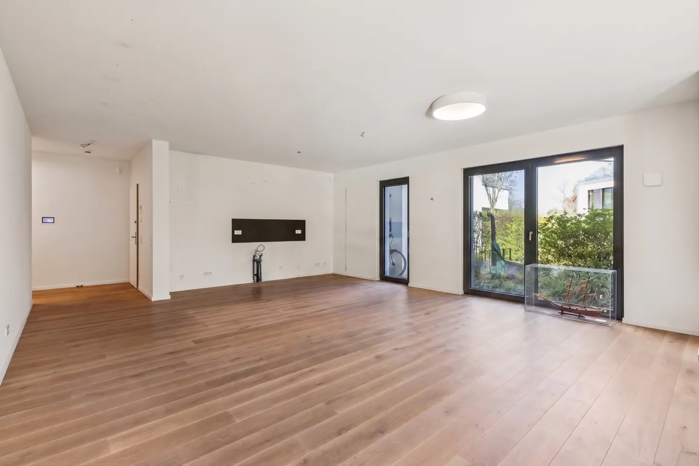 Bright, empty room with hardwood floors, white walls, and black-framed glass doors to the outside. A black panel is mounted on the wall.
