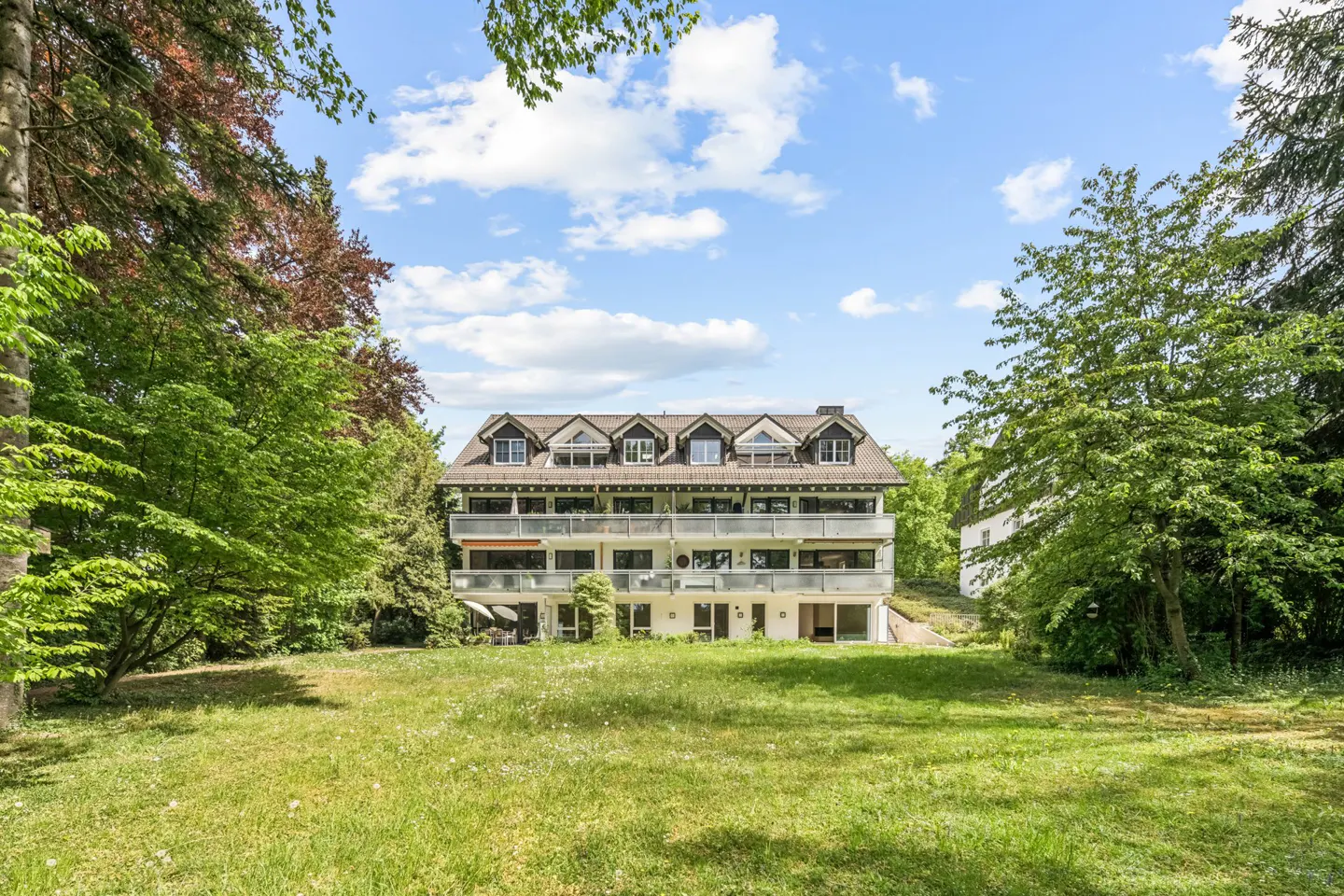 Exterior of a three-story white apartment building with balconies, surrounded by green trees and a grassy lawn under a blue sky.