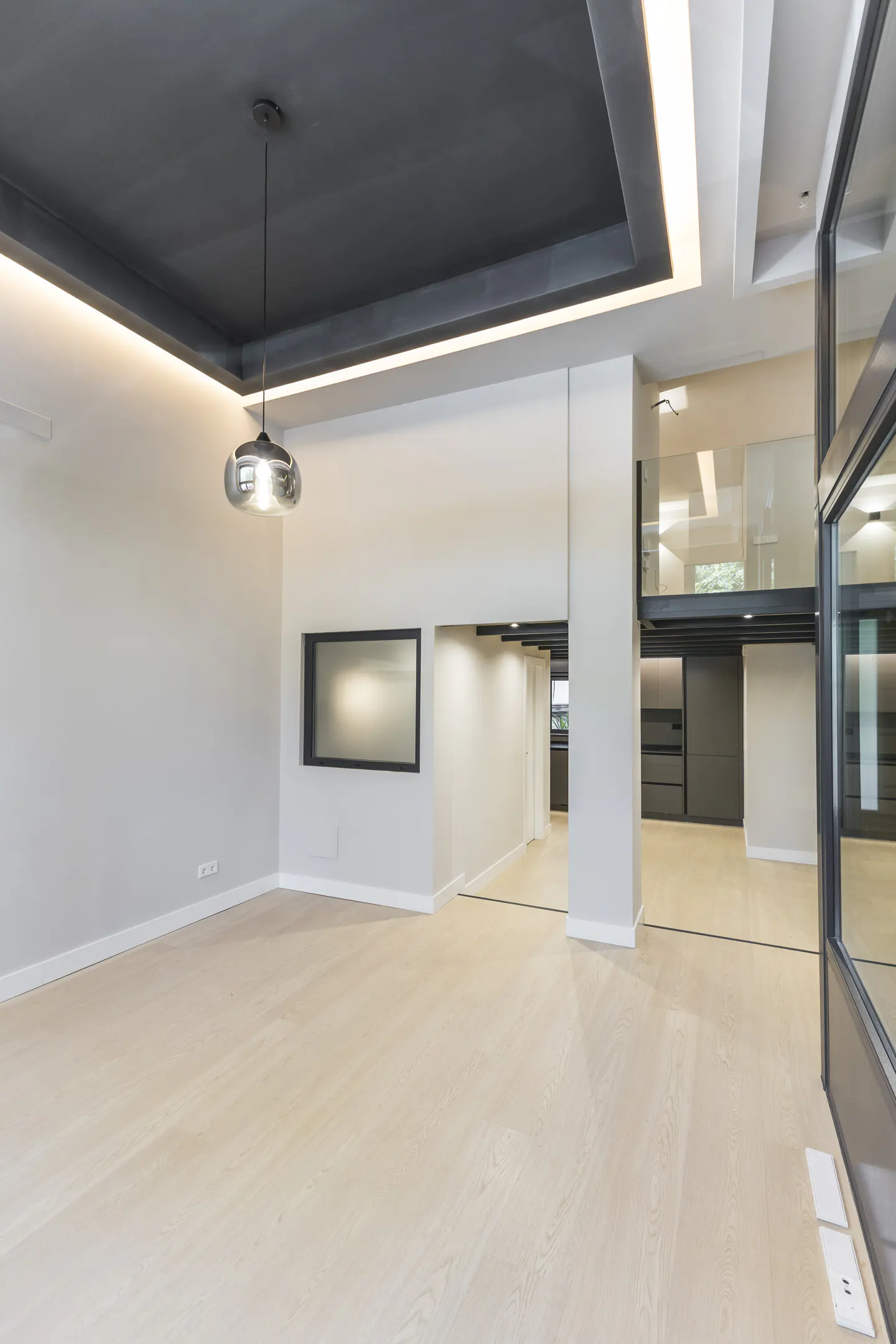 Modern open-plan living area with light wood floors, a dark tray ceiling with recessed lighting, and a glass mezzanine.