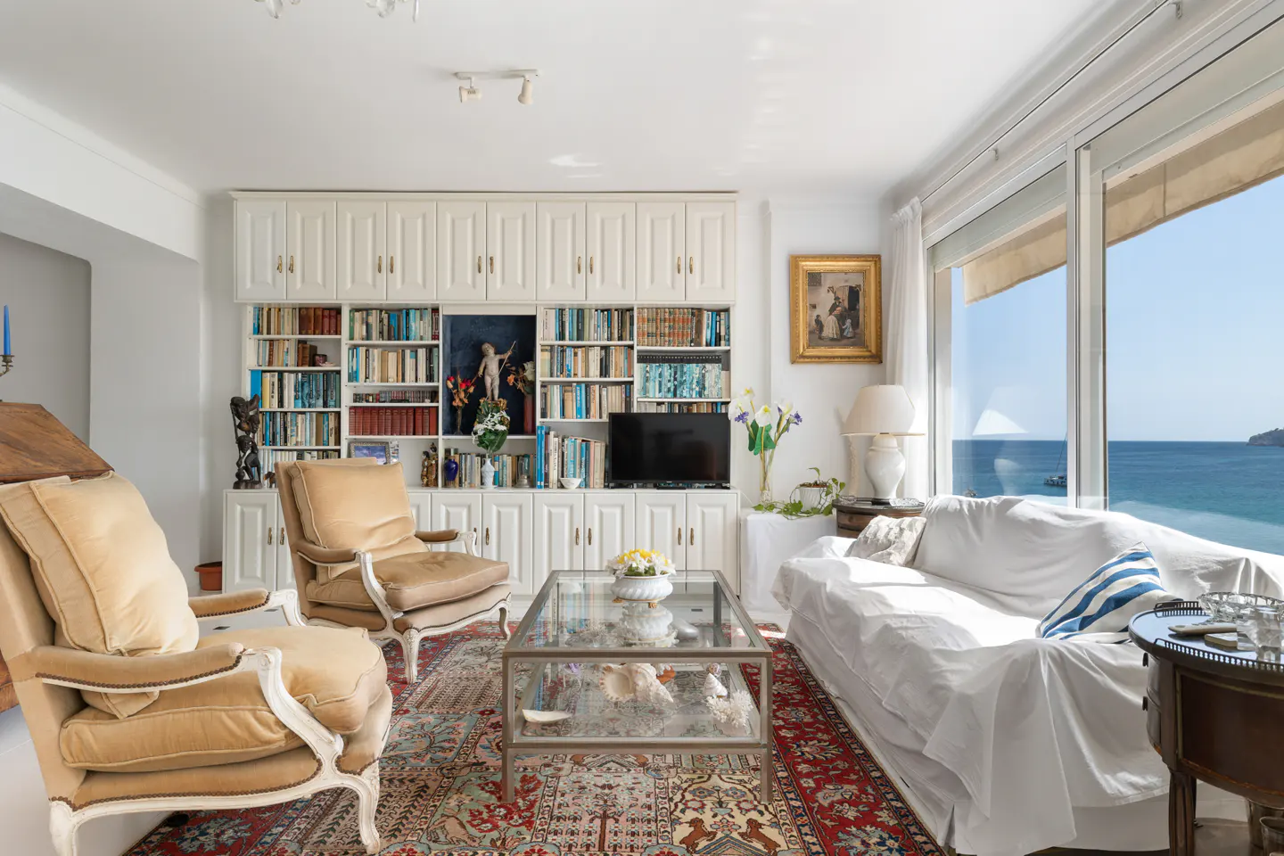 Living room with tan armchairs, white sofa, and a glass coffee table on a red patterned rug. Built-in bookcase and ocean view.