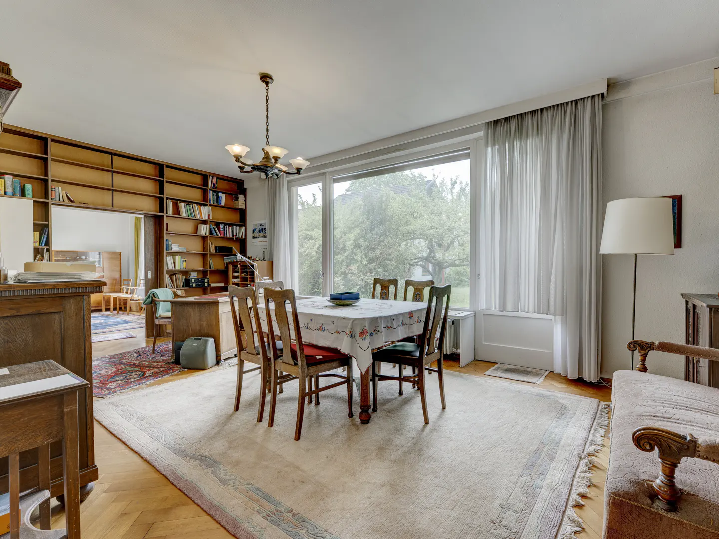 Dining room with a wooden table, chairs, large window with white curtains, floor-to-ceiling bookshelves, and area rug.