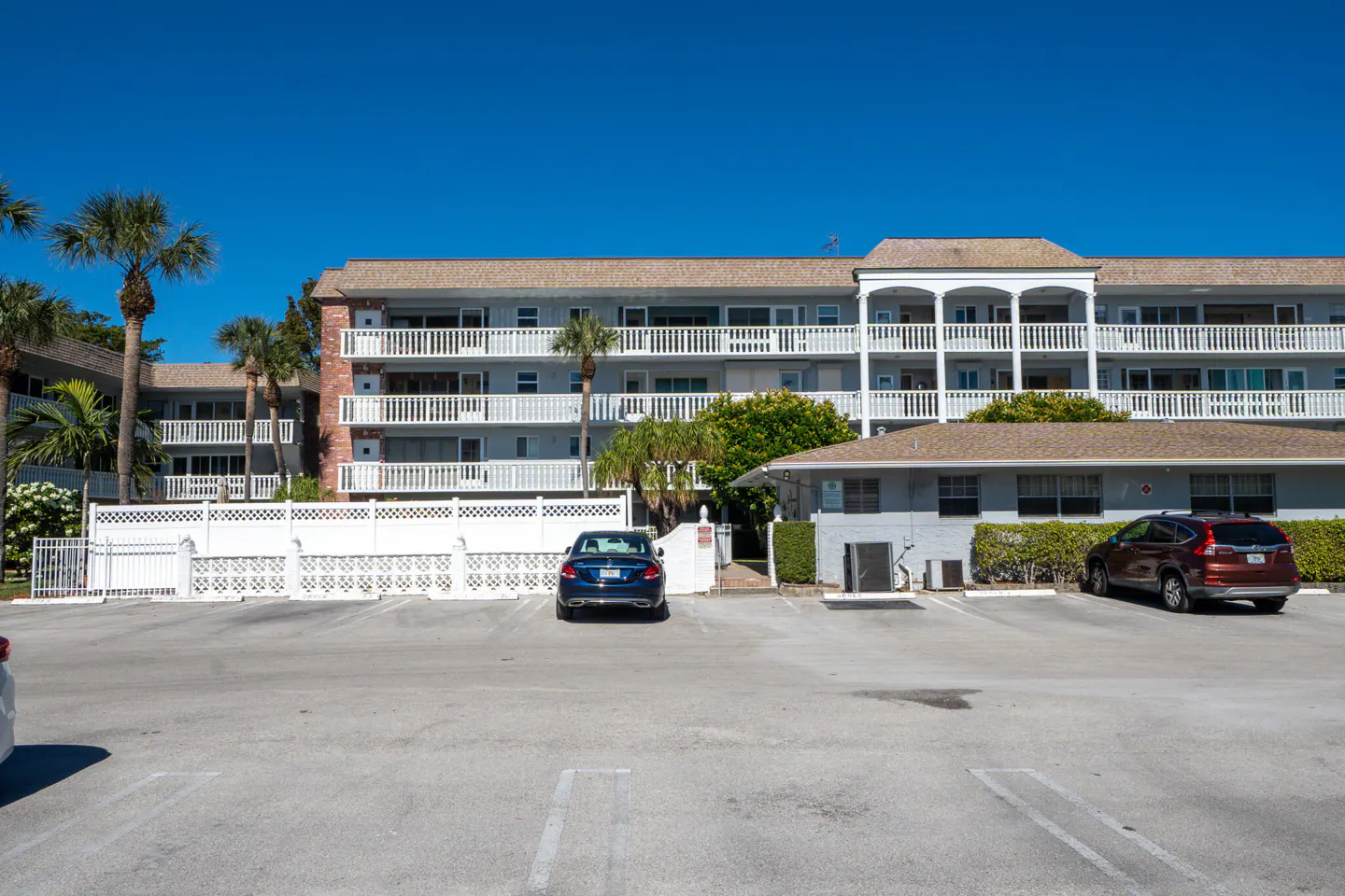 Exterior of a light gray condo building with white balconies and a parking lot with cars.