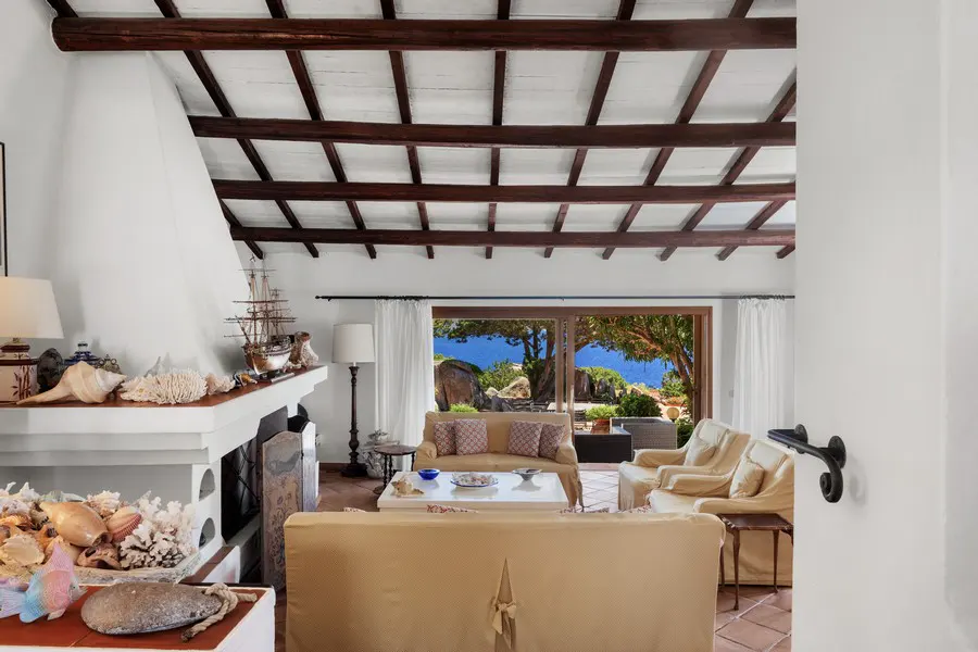 Living room with white walls, exposed beam ceiling, fireplace with shells, and ocean view through sliding glass doors.