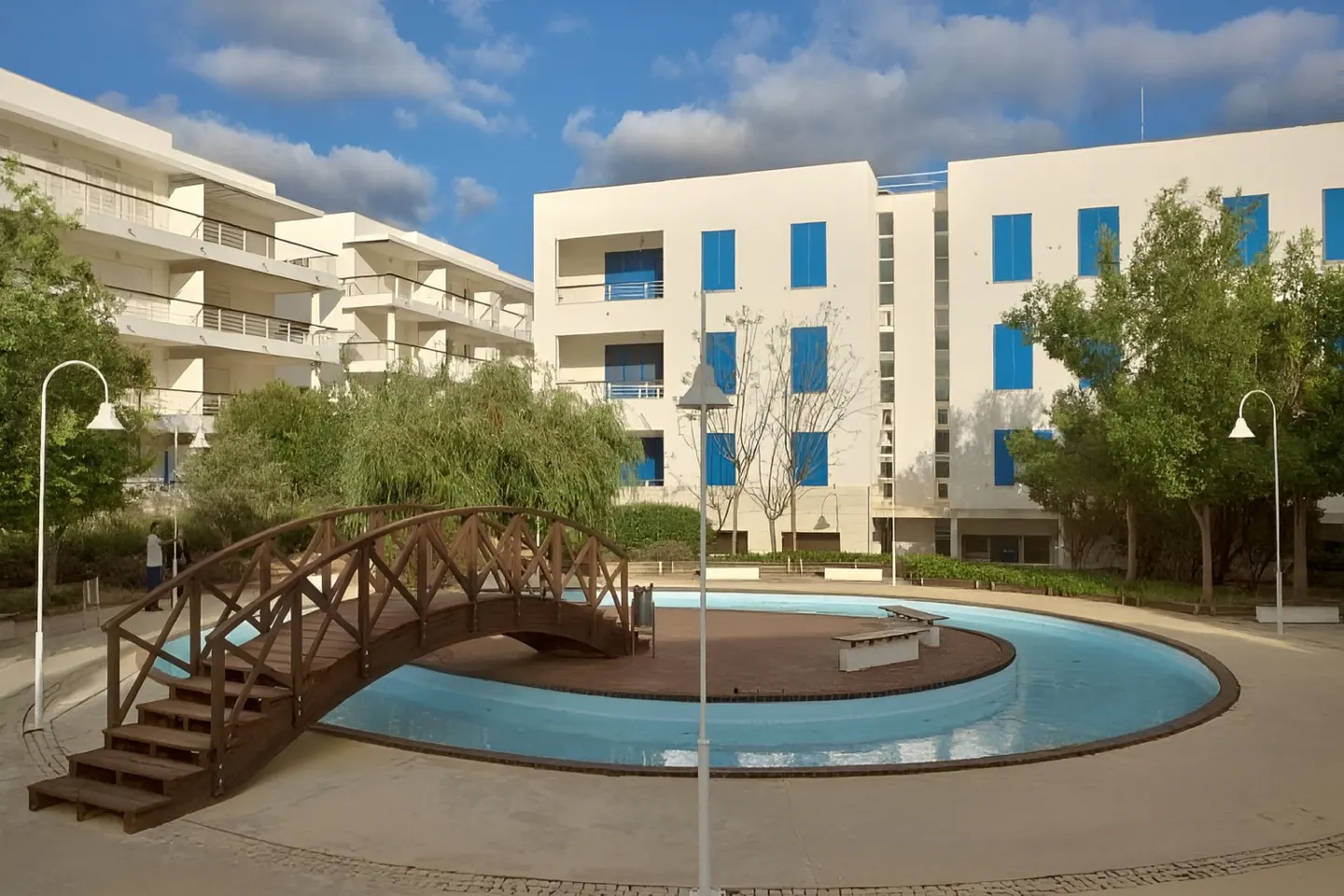 Exterior view of white apartment buildings with blue shutters, a wooden bridge over a water feature, and trees.