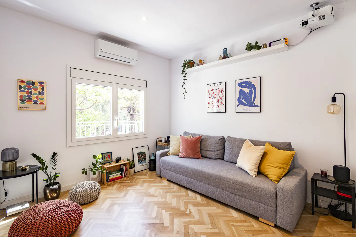 Bright living room with a gray sofa, colorful pillows, art prints, and herringbone wood floors. A projector is mounted on the ceiling.