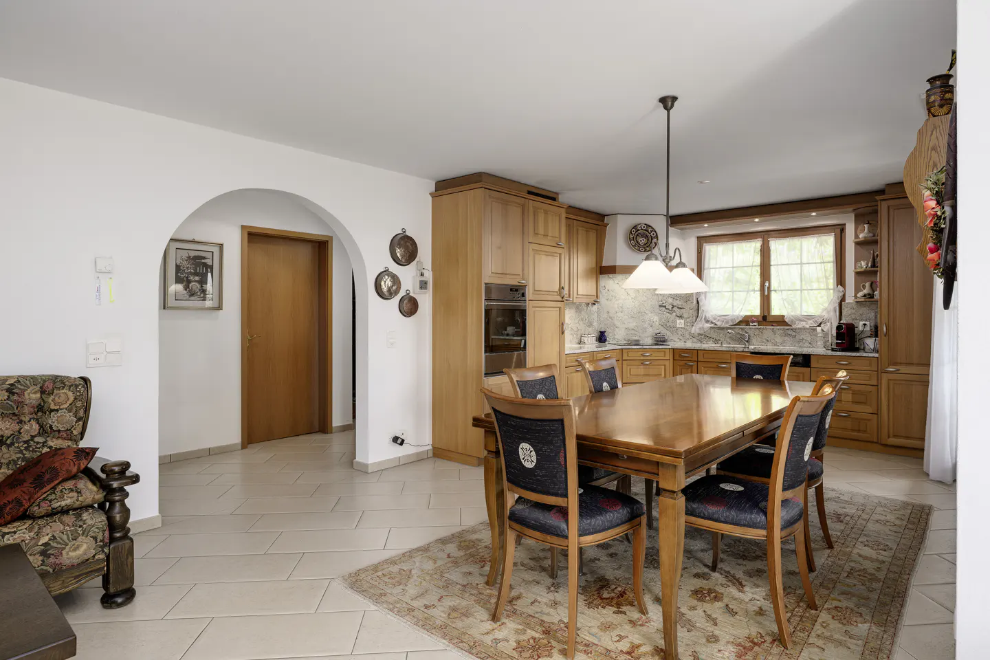 A bright, open-concept kitchen and dining area with wooden cabinets, table, and chairs. A patterned rug sits under the table.