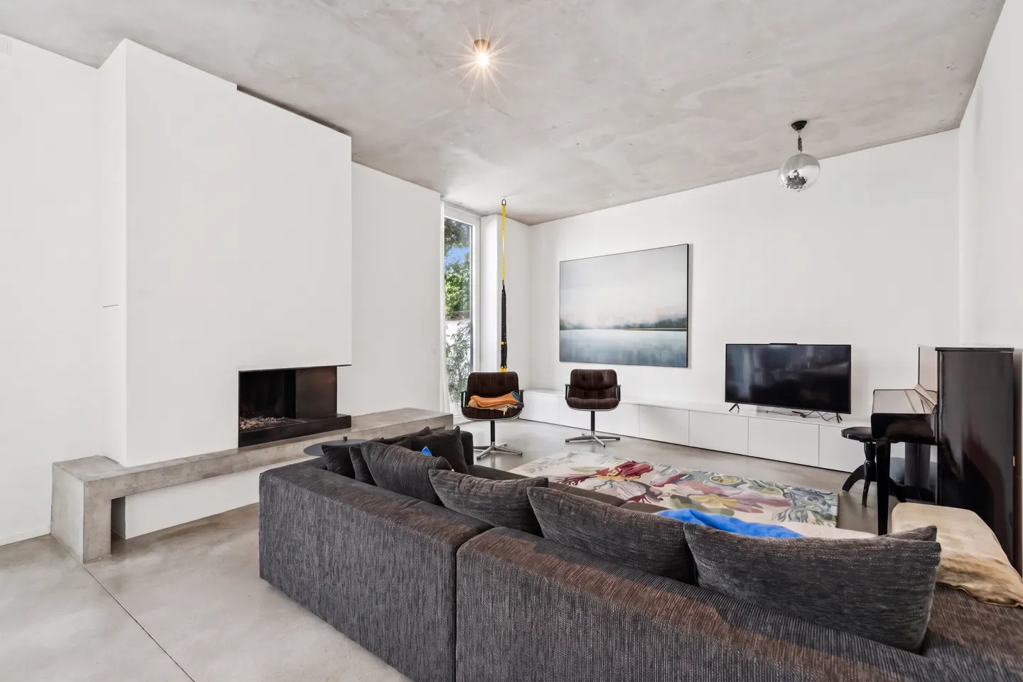 Bright living room with a gray sectional sofa, fireplace, two chairs, a piano, and a TV. A colorful rug adds a vibrant touch.