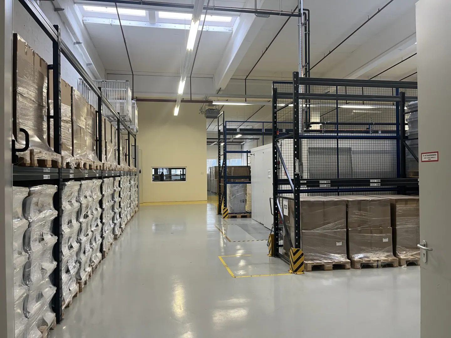A warehouse interior with shelves stocked with wrapped goods, a gray floor, and bright overhead lighting.