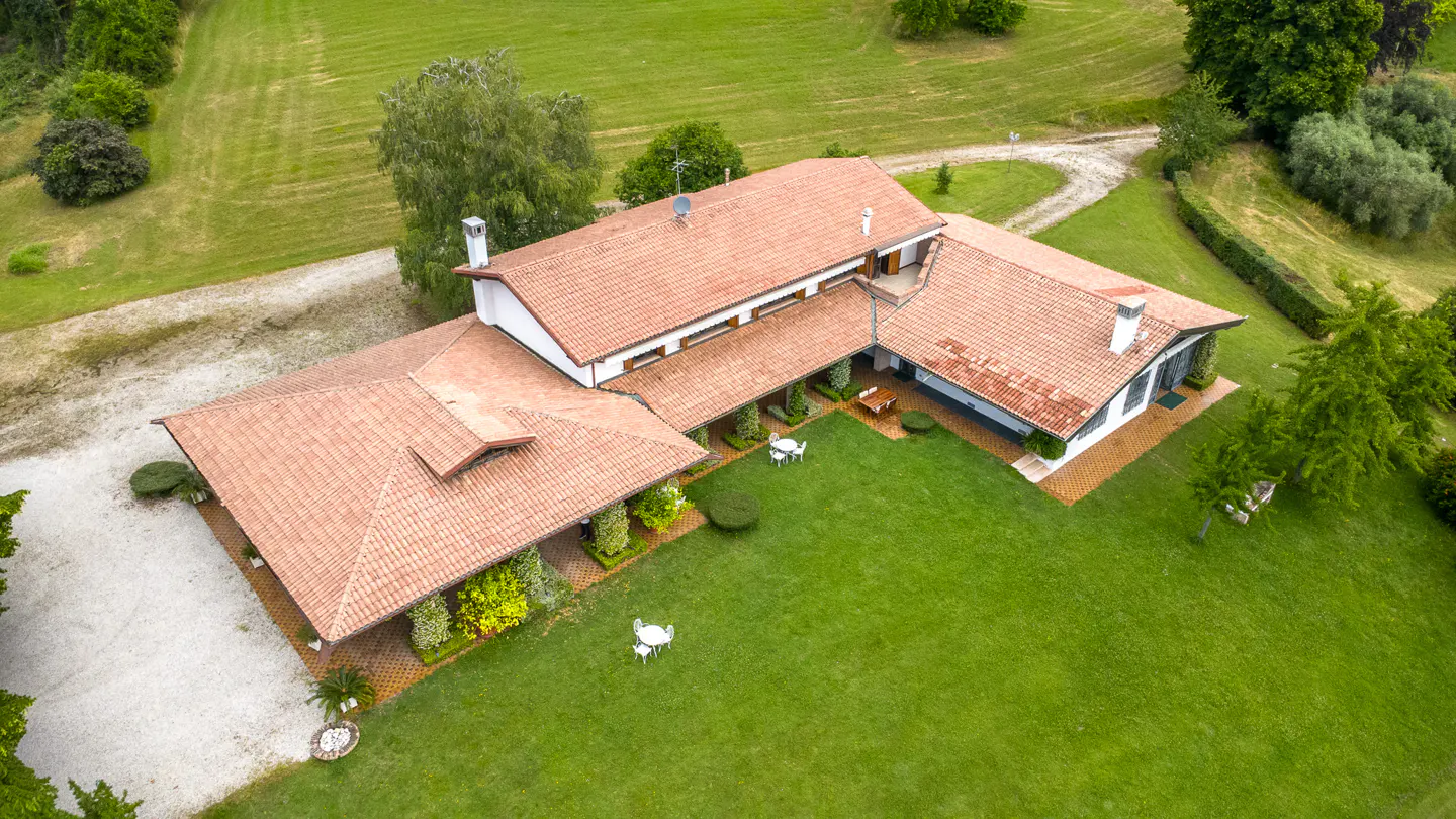 Aerial view of a white house with a terracotta tile roof, surrounded by a large green lawn and trees in a rural setting.