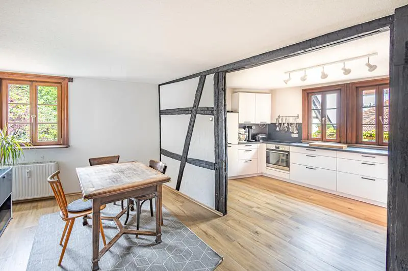 A bright kitchen and dining area with wood floors, white walls, and black timber framing. A wooden table and chairs sit on a gray rug.