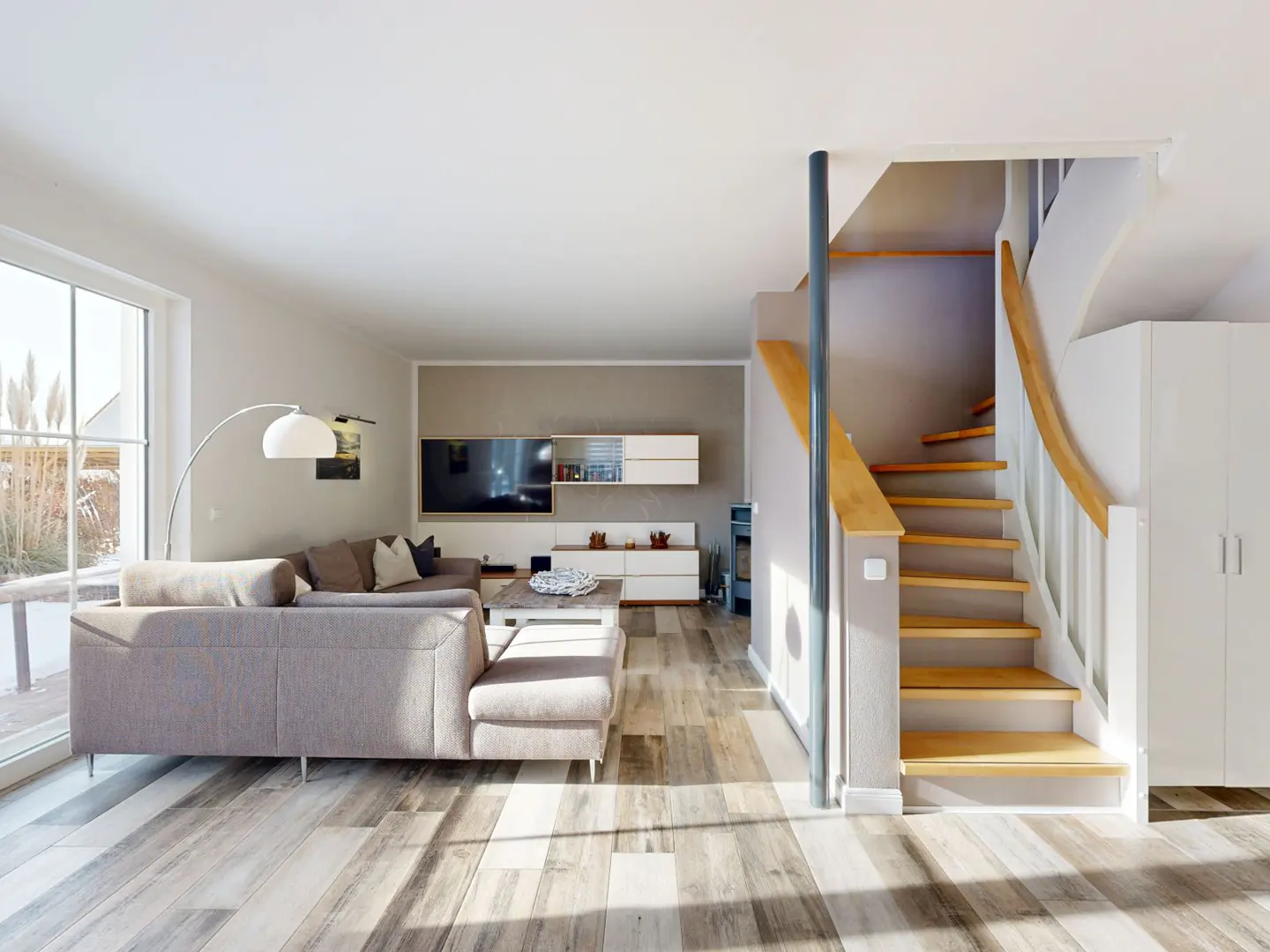 Bright living room with gray sectional sofa, wood floors, TV, and staircase with wood steps and white railing.