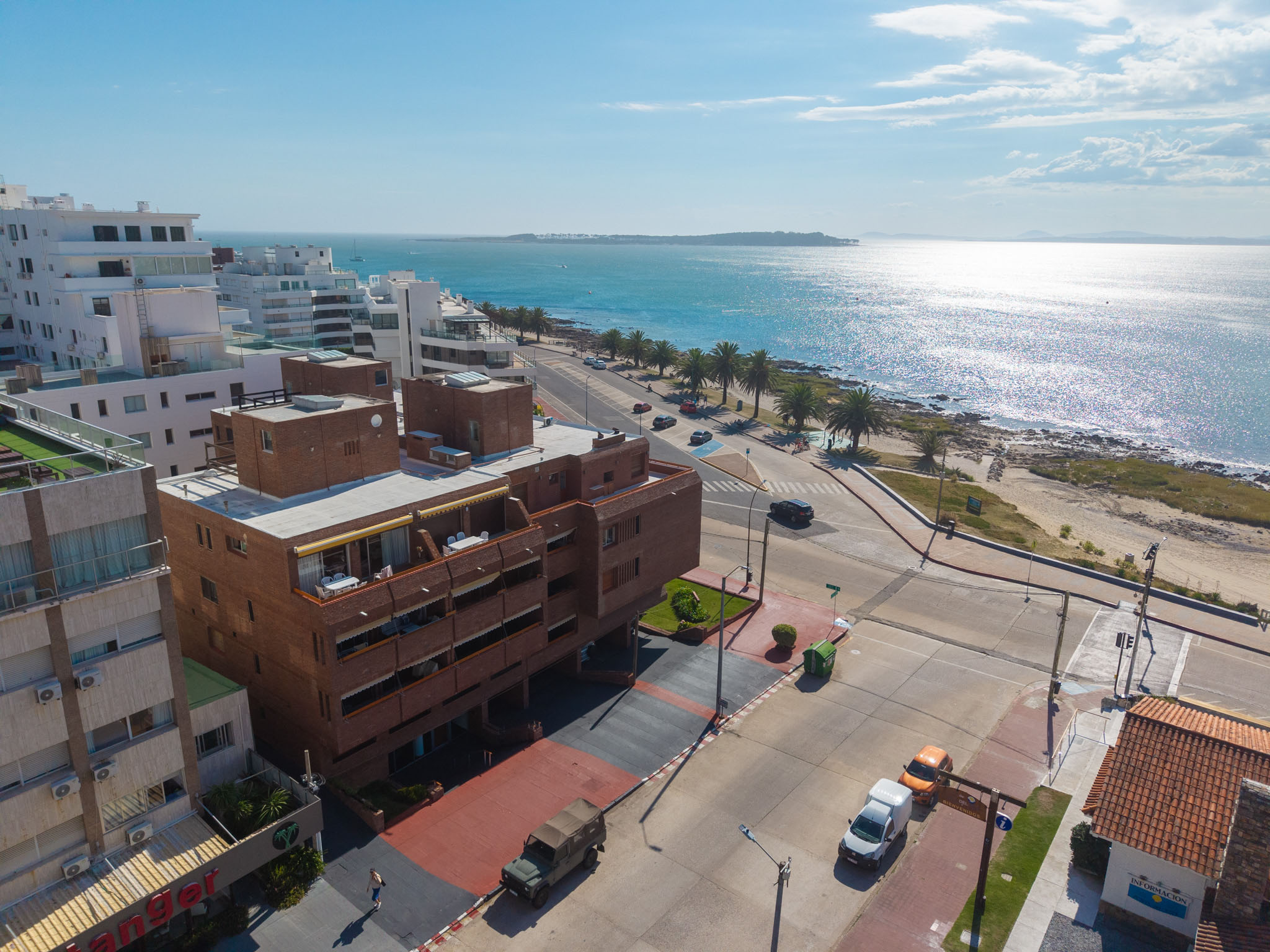 Aerial view of a coastal city with white buildings, a brown apartment complex, and a sparkling blue ocean under a sunny sky.