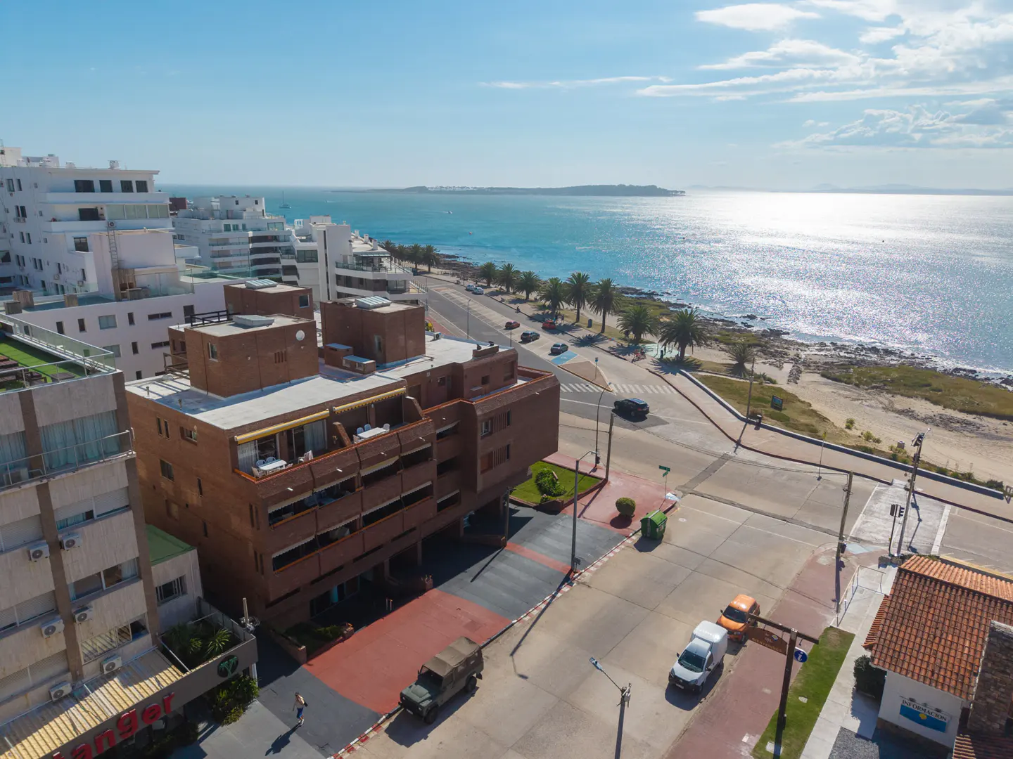 Aerial view of a coastal city with white buildings, a brown apartment complex, and a sparkling blue ocean under a sunny sky.
