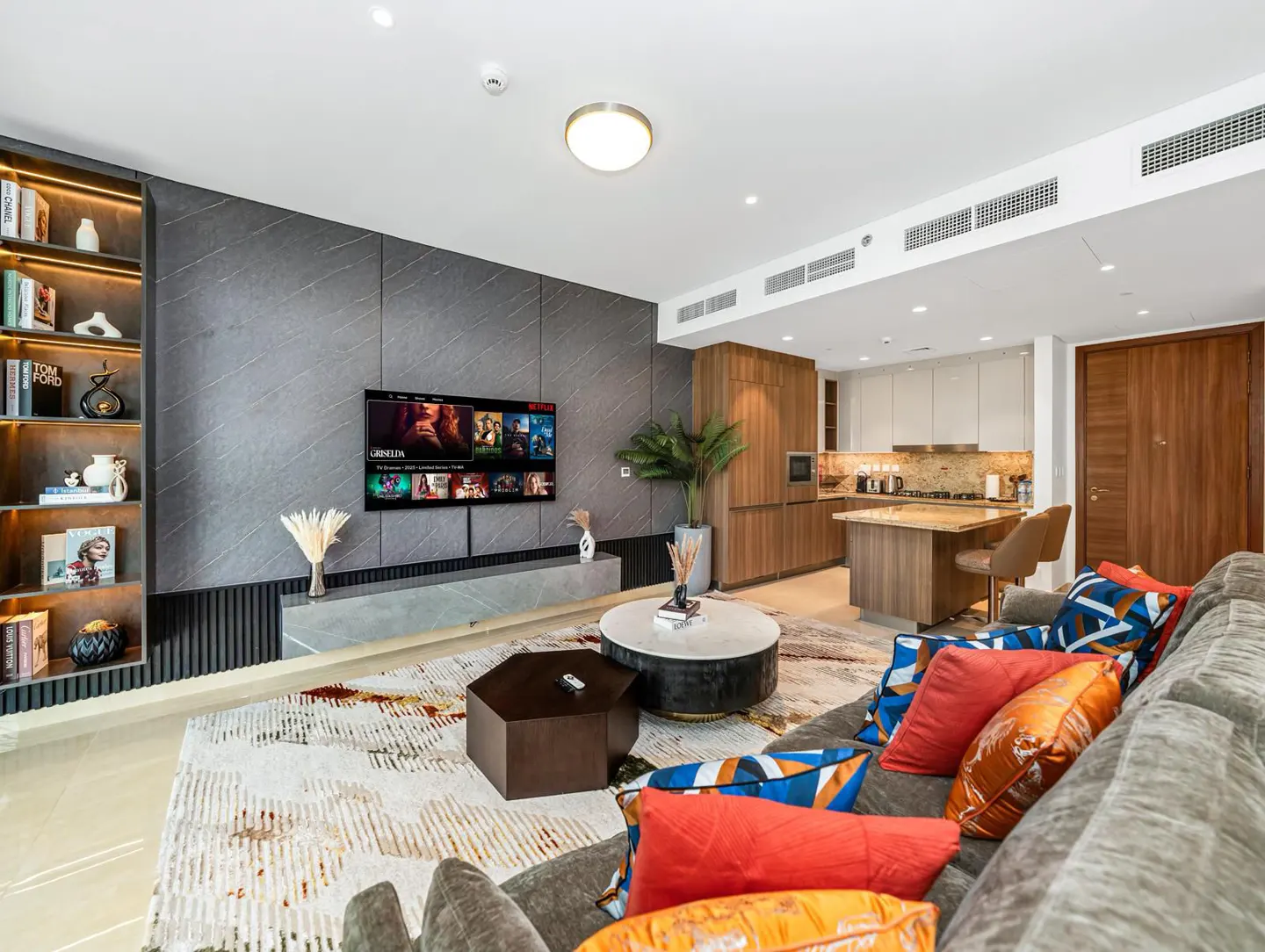 Open-concept living room with gray sofa, colorful pillows, round coffee table, TV, and kitchen in the background.