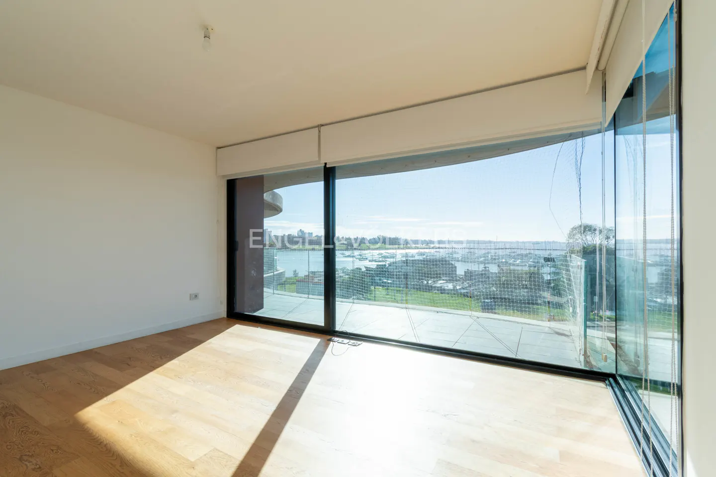 Bright, empty room with light wood floors and a large window overlooking a city and harbor.