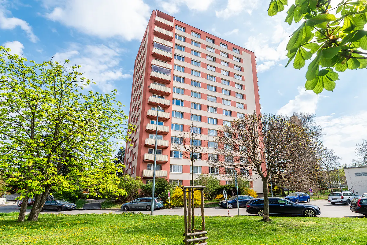 Exterior view of a tall, red and white apartment building with balconies, surrounded by green trees and parked cars.