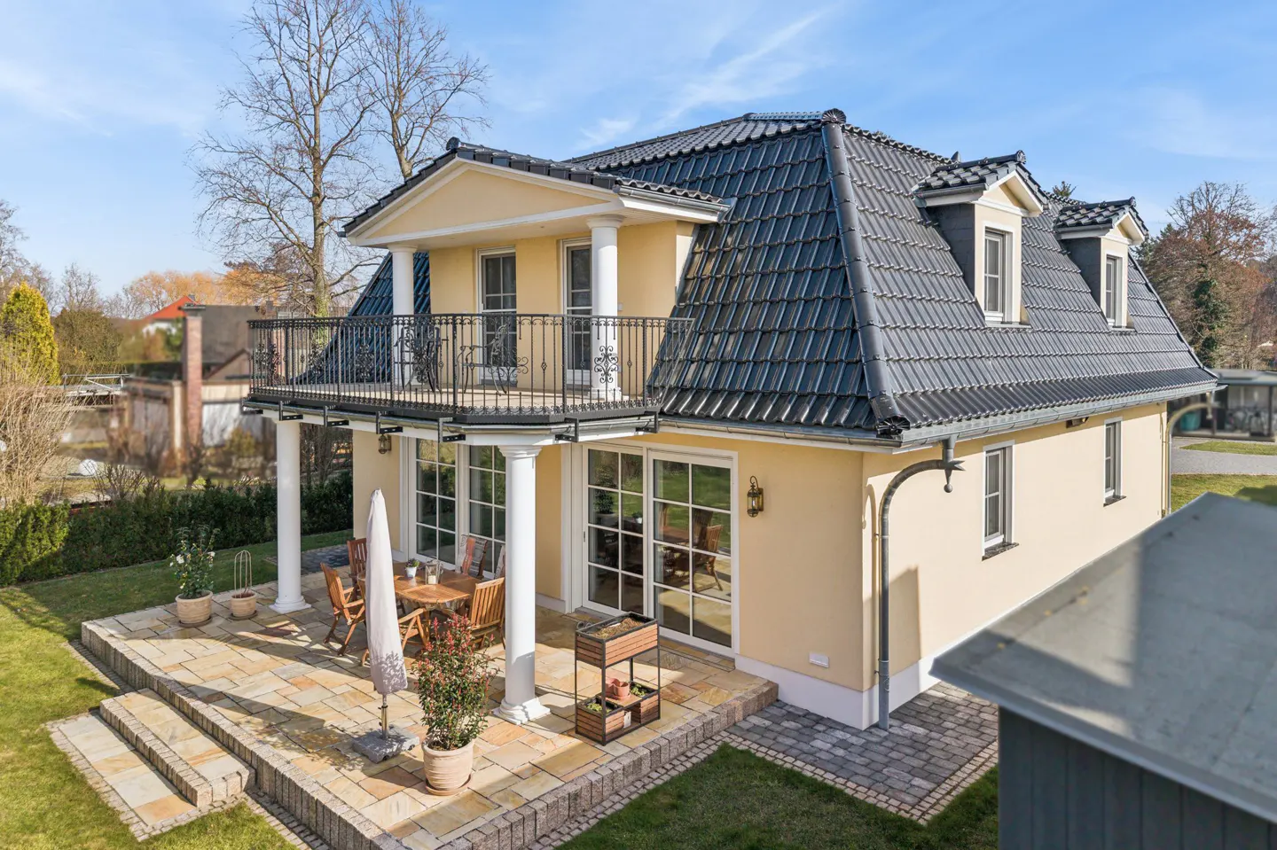 Two-story yellow house with a black tiled roof, balcony, patio, and lawn on a sunny day.
