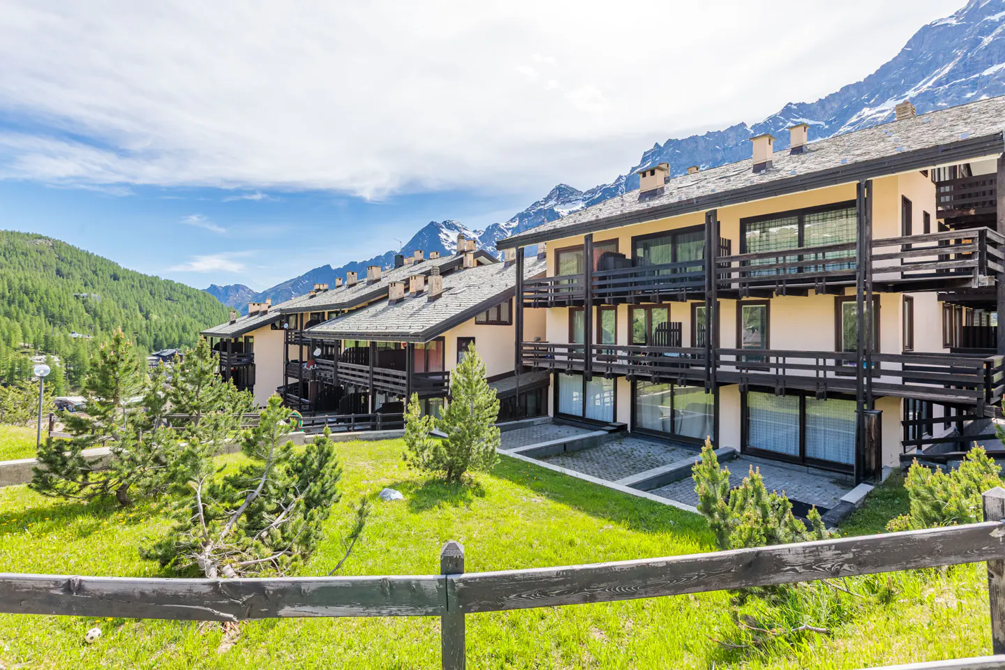 Multi-story apartment building with wooden balconies set against a backdrop of snow-capped mountains and green hills.