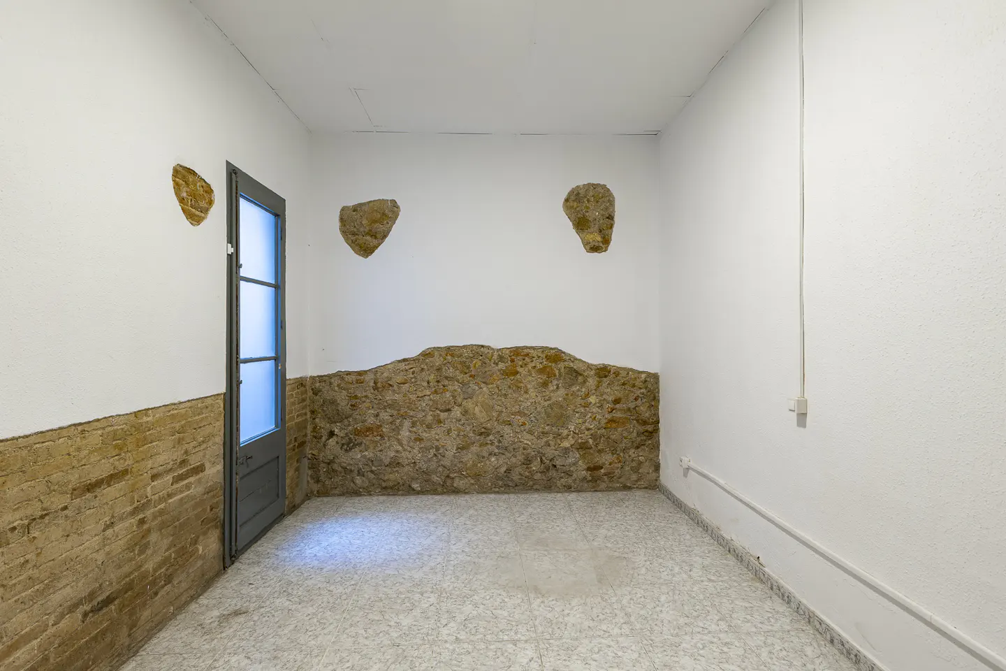 Empty room with white walls, brick accents, and a gray door. The floor is tiled in a light pattern.