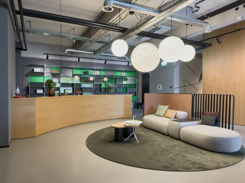 Modern office lobby with a curved wooden reception desk, colorful shelving, and a gray modular sofa on a round rug. Overhead lighting fixtures.