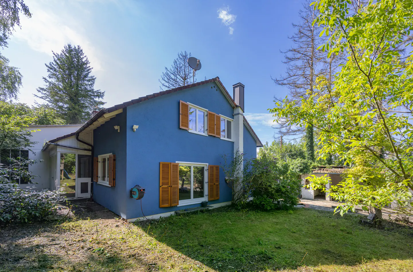 A two-story blue house with brown shutters and a red roof, surrounded by green trees and a lawn.